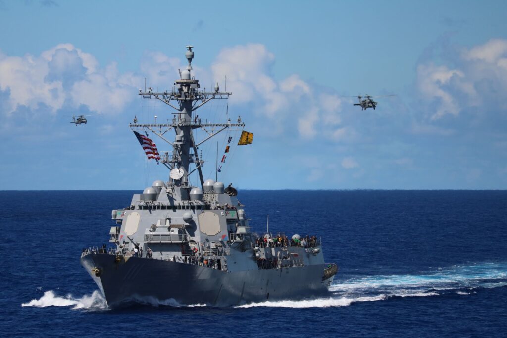 A US Navy destroyer navigates the ocean with helicopters in formation above.