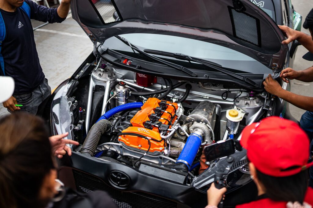A group inspecting a car engine at an outdoor automative event, hood open.