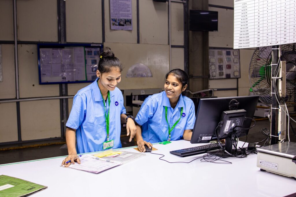 Two female workers discussing tasks at a factory workbench, smiling and engaged.