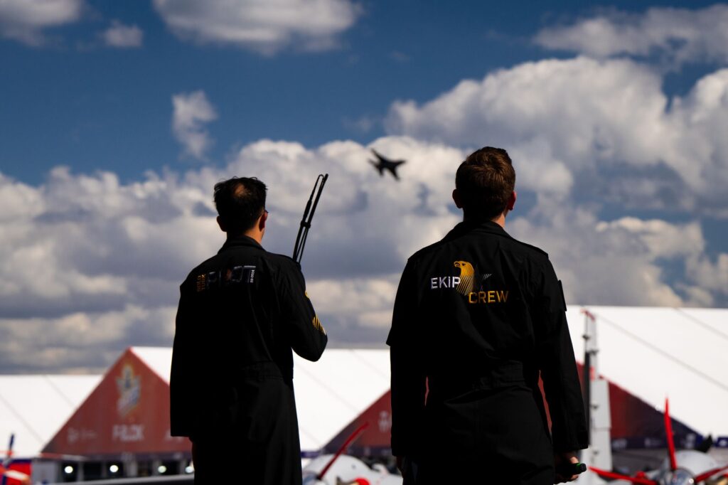 Two crew members observe a jet performing in the sky at an airshow under clear conditions.