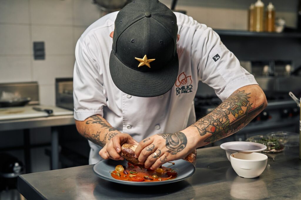 Tattooed chef expertly plates a gourmet dish with precision and skill in a restaurant kitchen.