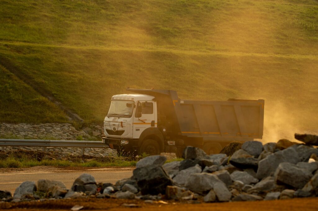 A dump truck drives along a dusty road, illuminated by golden sunlight, with rocks visible in the foreground.