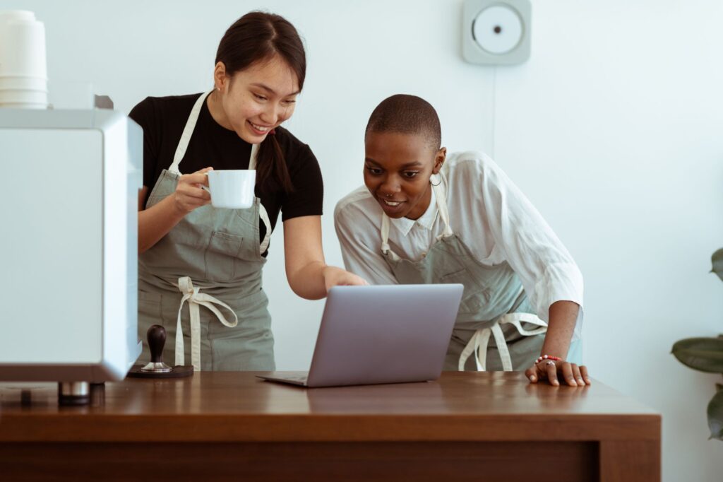 Positive smiling young multiracial waitresses wearing aprons gathering near table with cup of hor drink and surfing modern netbook in light kitchen