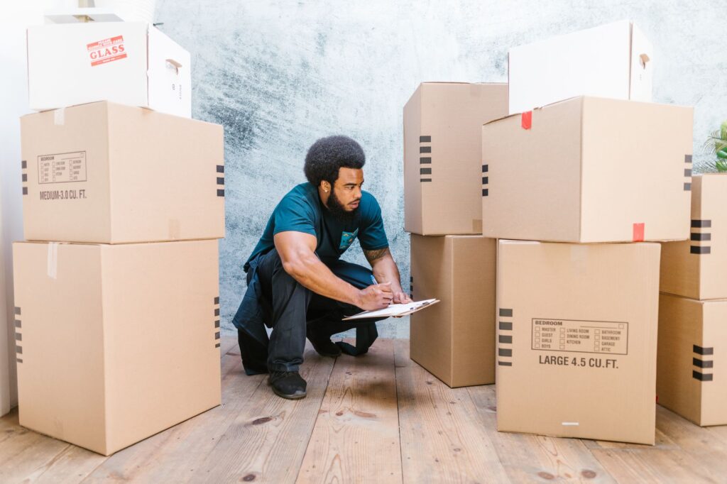 An adult man in workwear checks a clipboard while surrounded by moving boxes in an indoor setting.