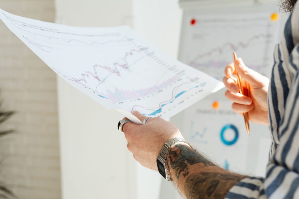 Close-up of a tattooed man's hands holding financial charts, with focus on data analysis and stock market trends.