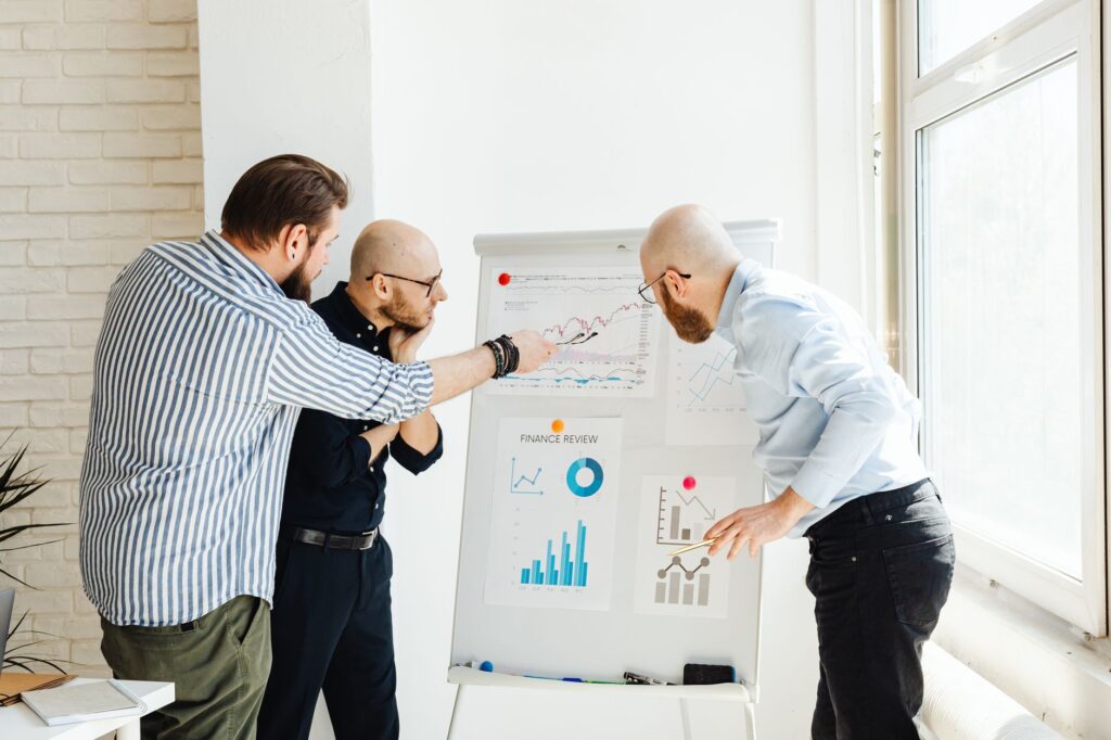 Three colleagues in a bright office discuss financial charts on a whiteboard.