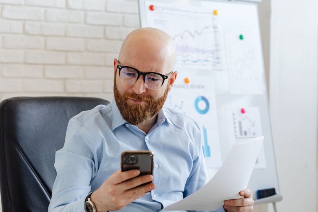 Bald businessman with eyeglasses checking phone data during an office presentation.